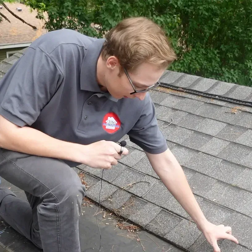 Man kneeling on a shingled roof, inspecting it with a tool near the edge.