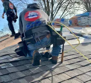 Two roofers kneel on a shingled roof, carrying bundles of shingles with safety harnesses attached.