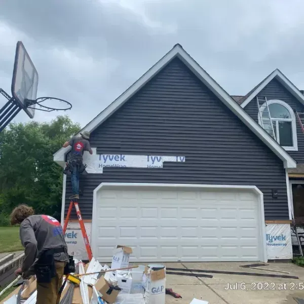Two workers install house siding and trim on a dark gray home with a white garage and basketball hoop.