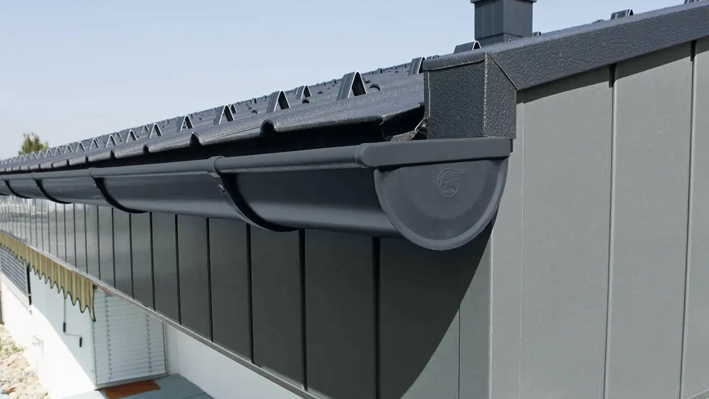 Black rain gutter along the edge of a gray building roof under a clear sky