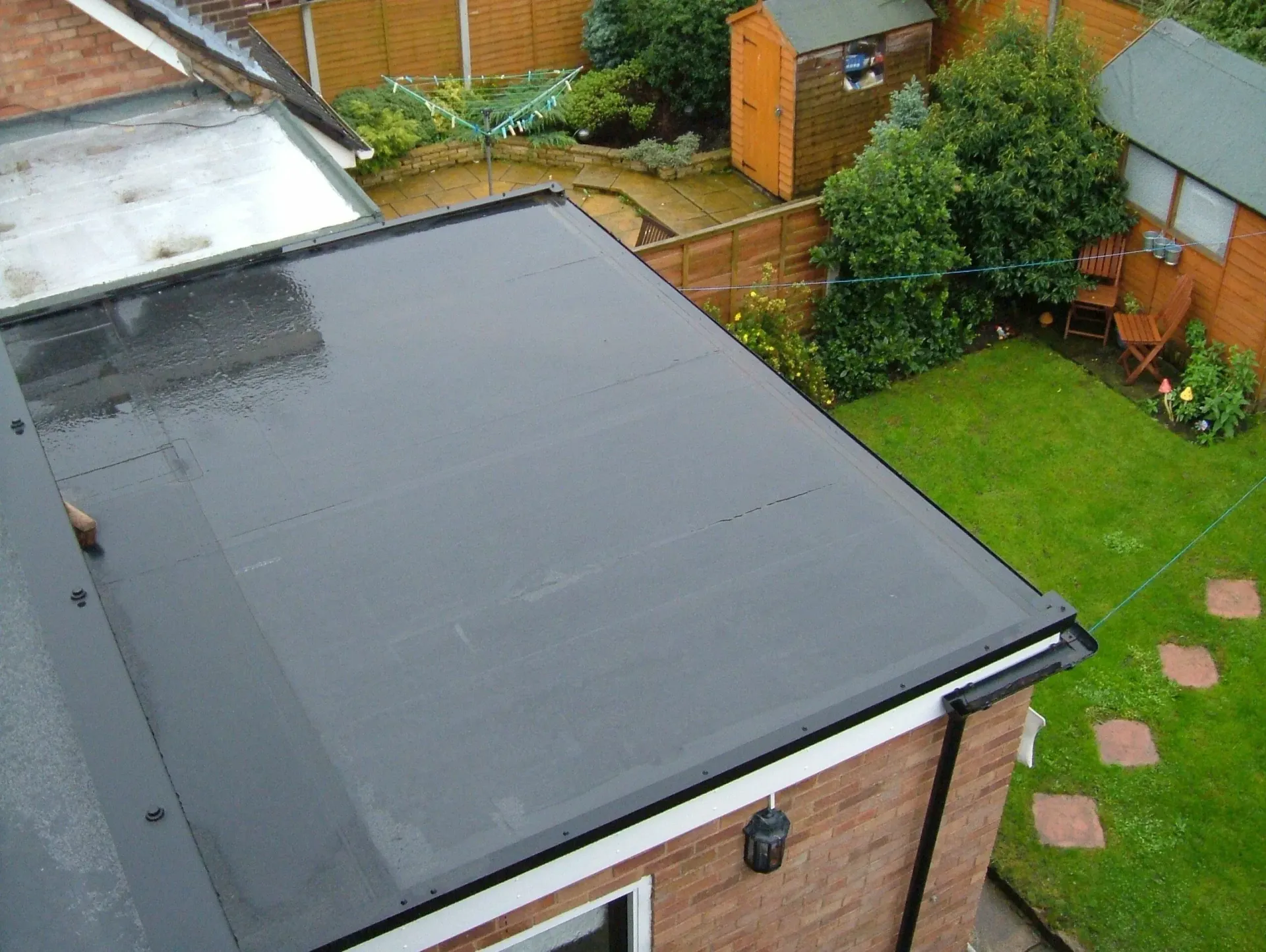 Roof of a house with a dark flat rooftop, surrounded by a small backyard with grass, fence, and sheds