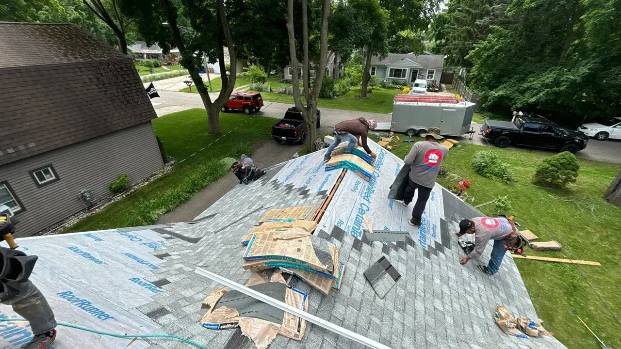 Roofers in safety gear work on a residential roof covered in underlayment, with houses and vehicles visible below.