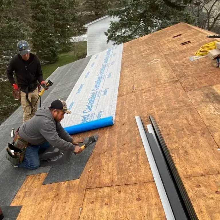 Workers installing roofing underlayment on a sloped roof with shingles and materials nearby