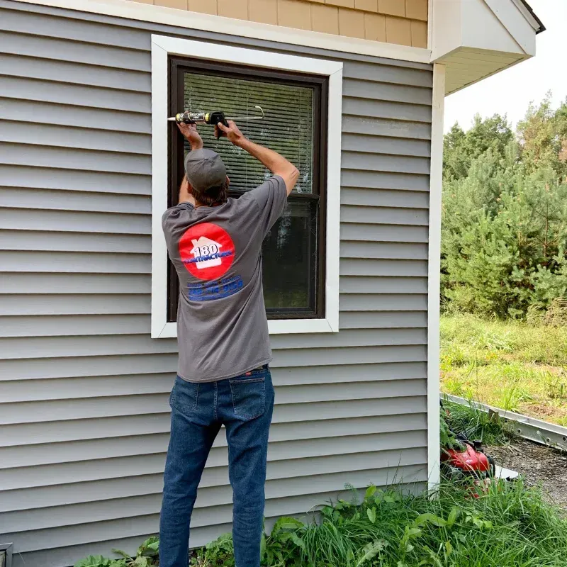 Worker installing or adjusting a window screen on a gray house exterior.