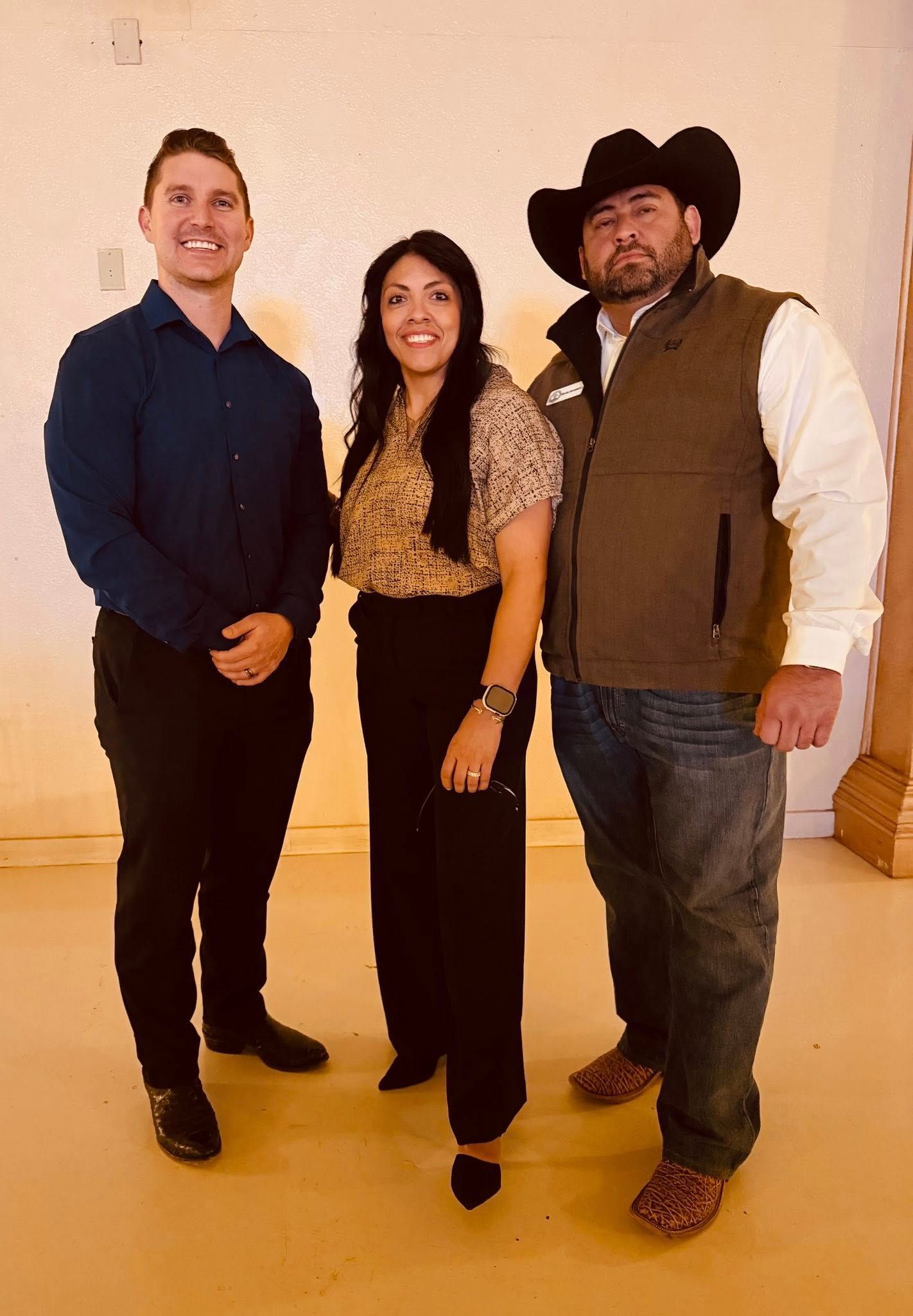 Three people posing indoors against a plain wall, smiling at the camera.