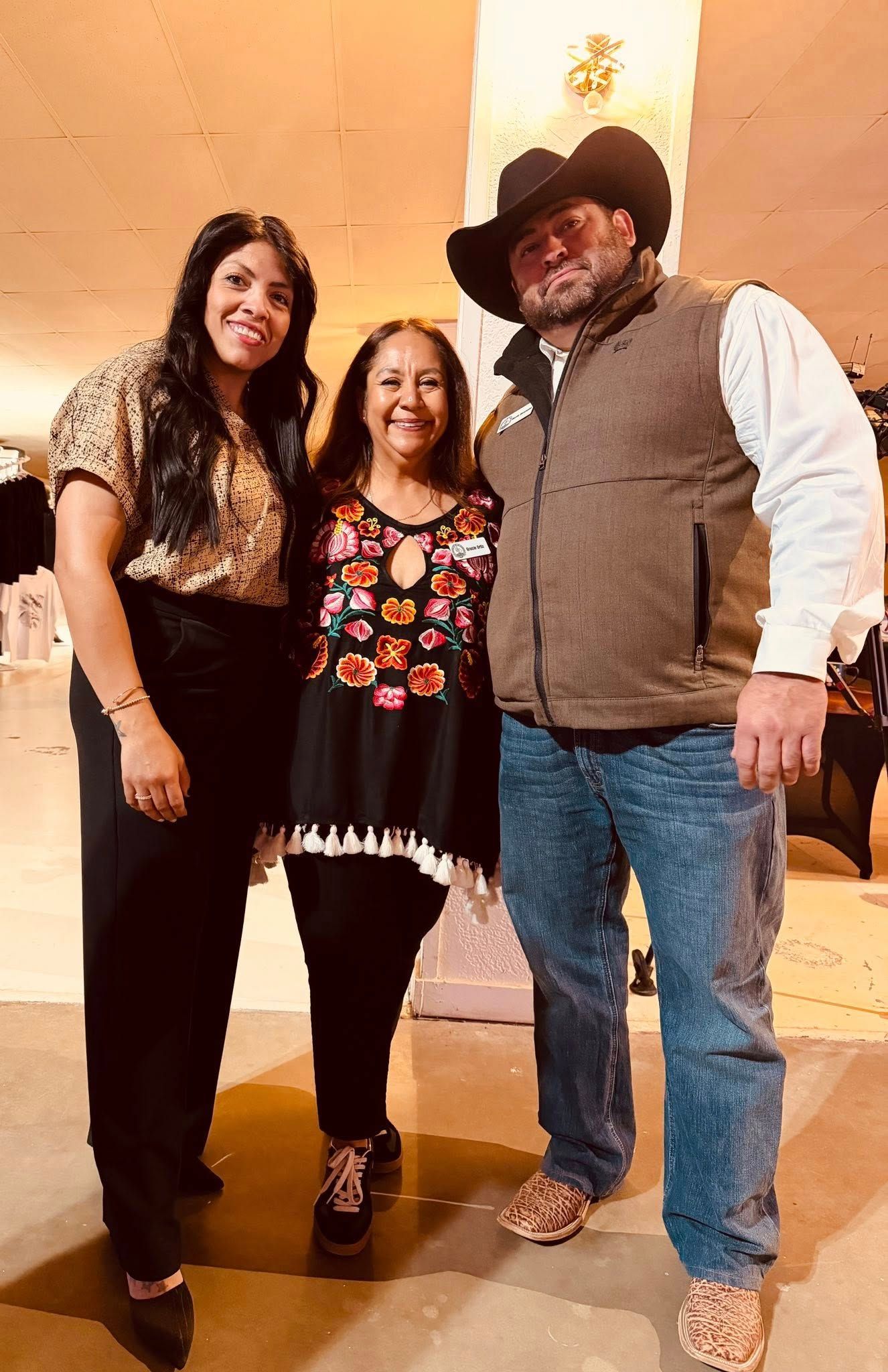 Three people posing together indoors, smiling in front of a warm beige wall.