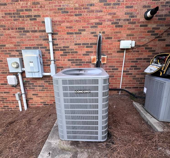 Outdoor HVAC unit beside a brick wall with utility boxes and pipes, next to another air conditioner unit.