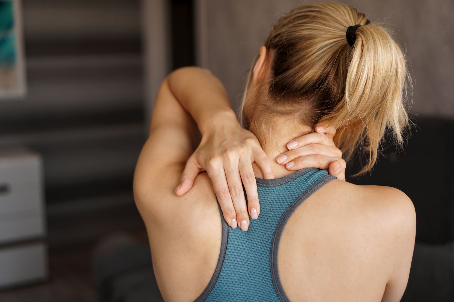Woman in teal tank top massaging her neck and shoulder indoors