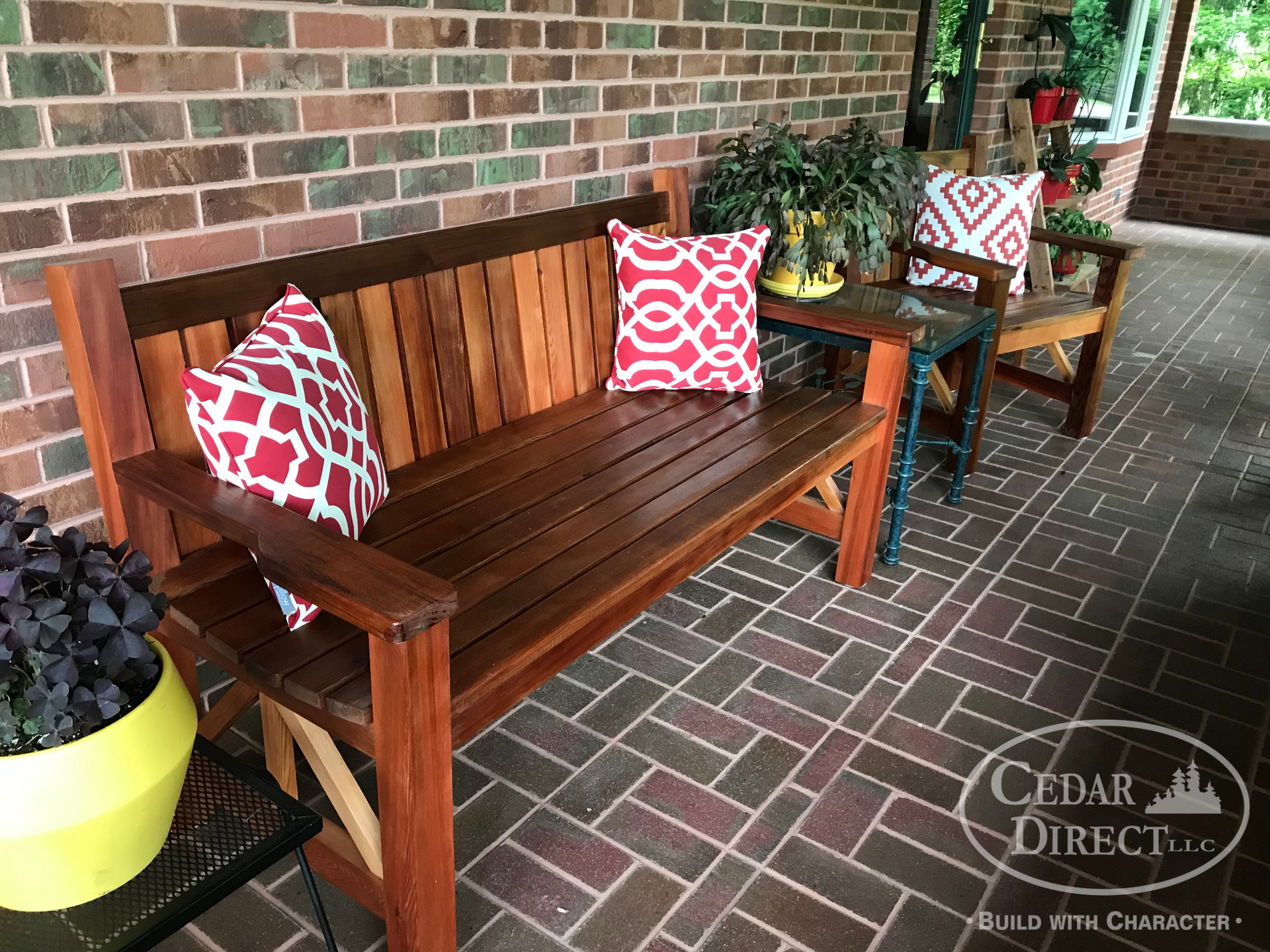 A wooden bench with red and white pillows on it is on a brick porch.