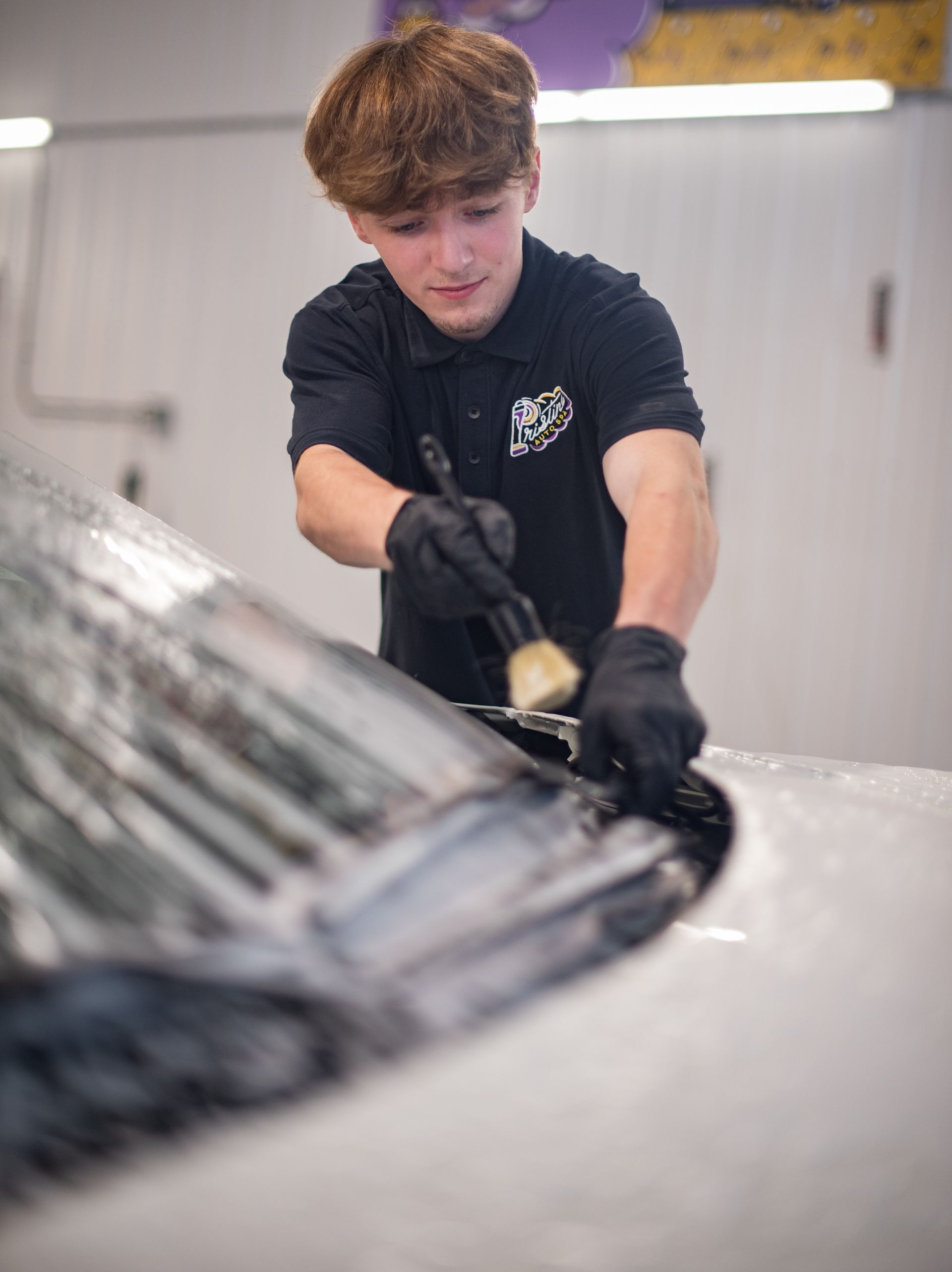 Person cleaning a car windshield with a brush, wearing a black shirt and gloves.