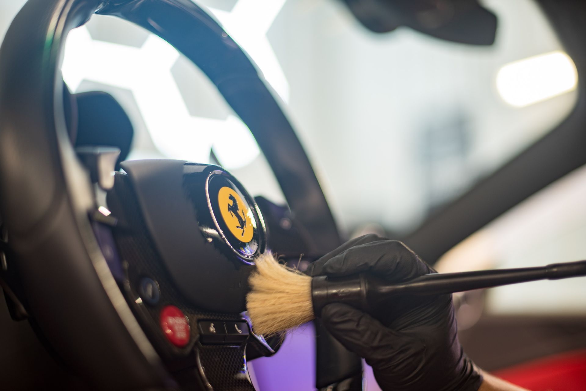 Gloved hand uses a brush to clean a Ferrari steering wheel emblem with a yellow prancing horse logo.