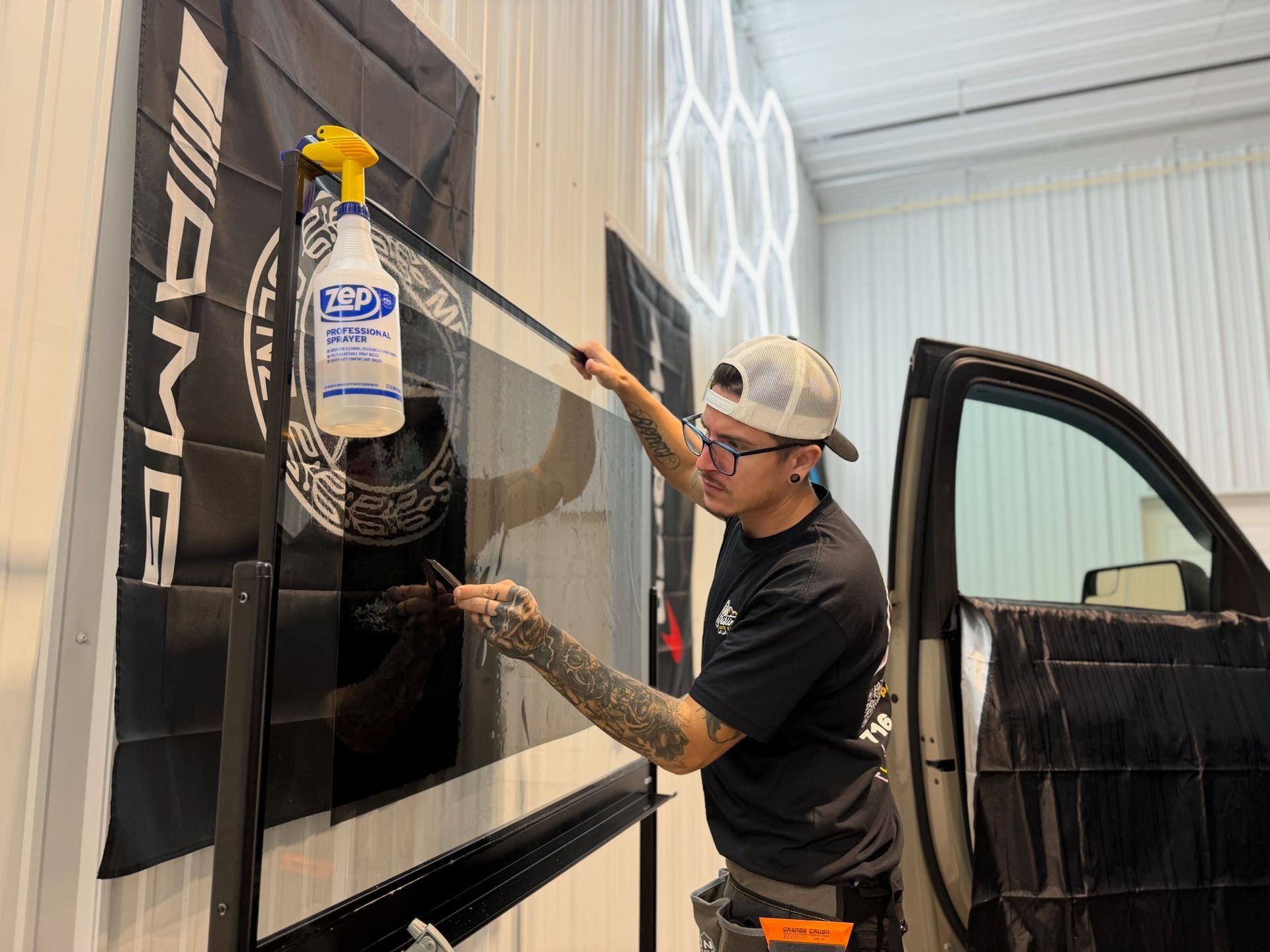 Man applying tint to car window, holding a spray bottle. AMG banner in background.