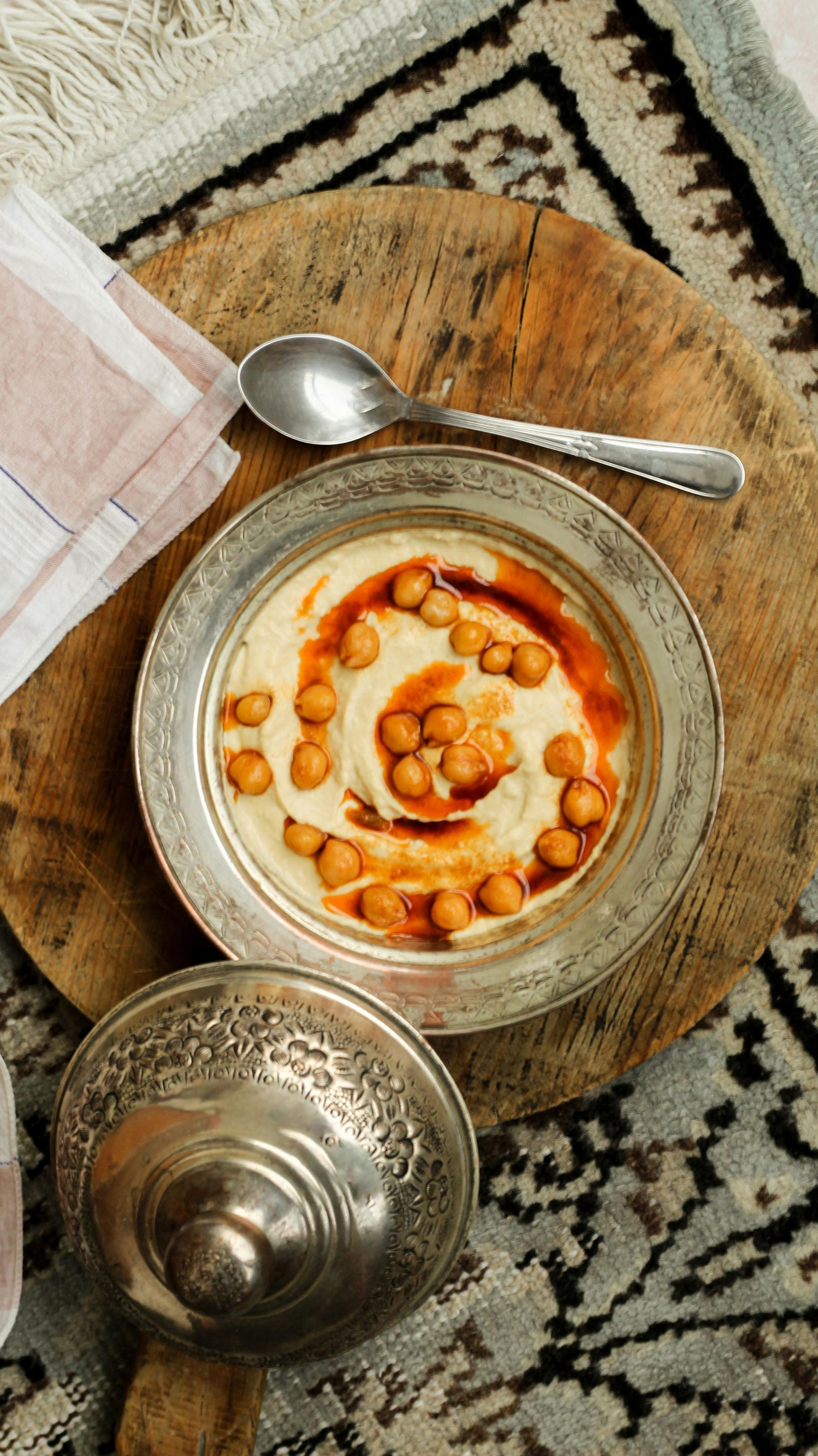 Een kom hummus met kikkererwten en een lepel op een houten tafel.