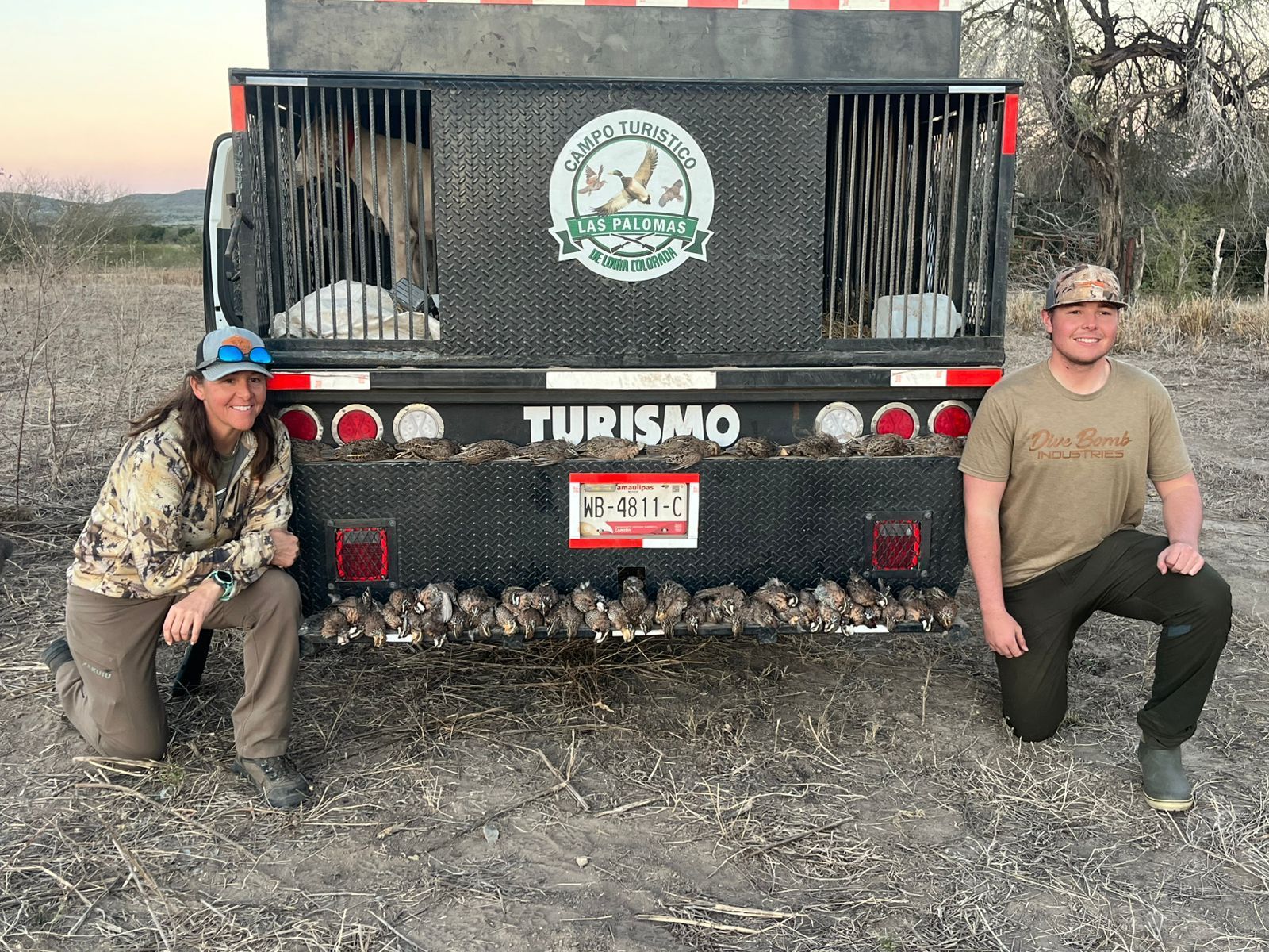 Two people crouch beside a black off-road truck with a front grille and “TURISMO” on the bumper.