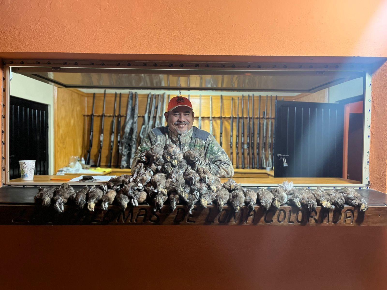 Smiling hunter behind a table lined with hanging ducks and rifles in a wooden duck blind