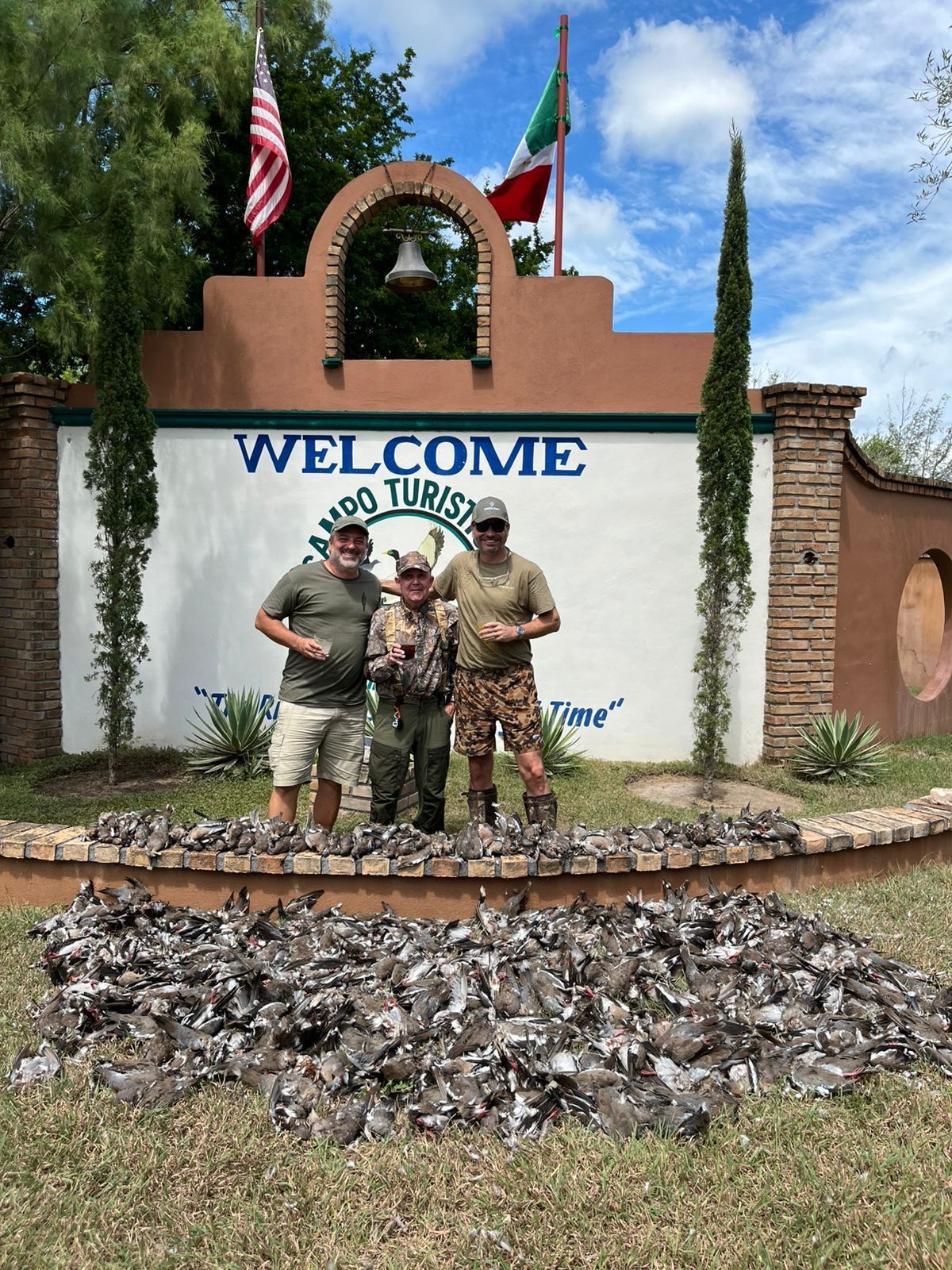 Two soldiers posing in front of a welcome sign and flags at a military-themed entrance monument.
