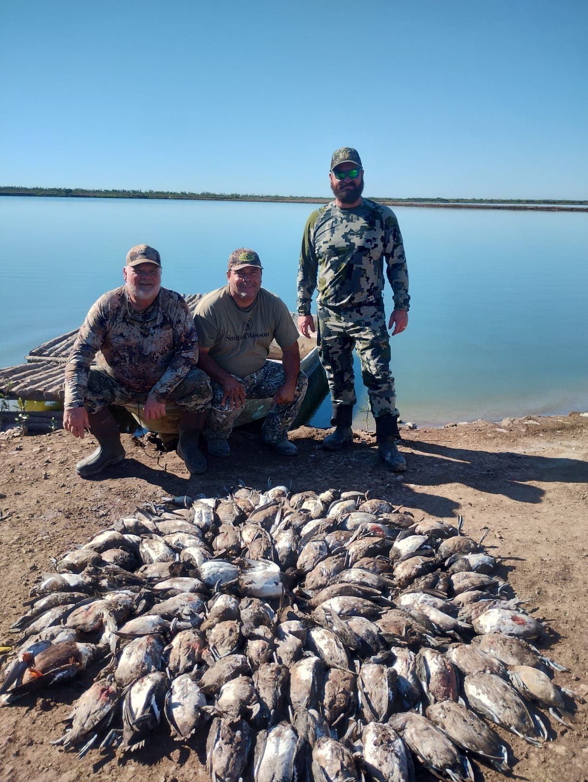 Three camouflaged people pose beside a large pile of fish on a rocky lakeshore.