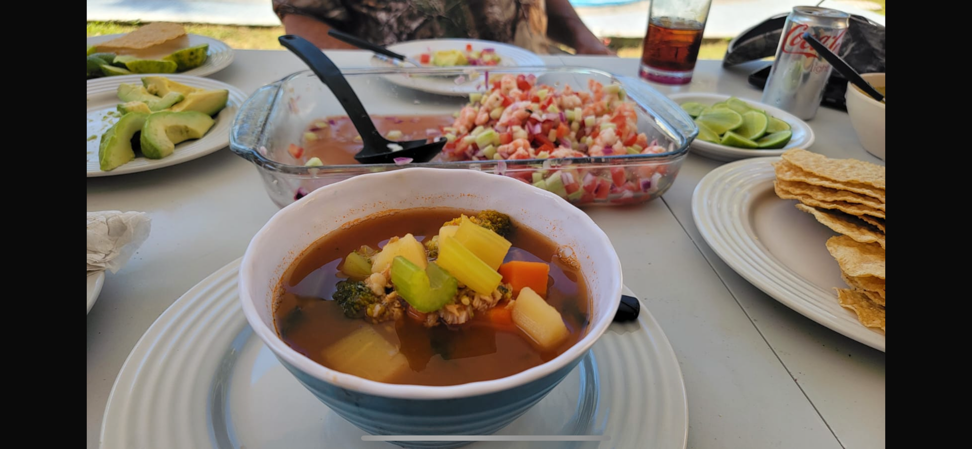 Table with soup, salad, avocado slices, and crackers on plates, with drinks and utensils in a casual dining setting