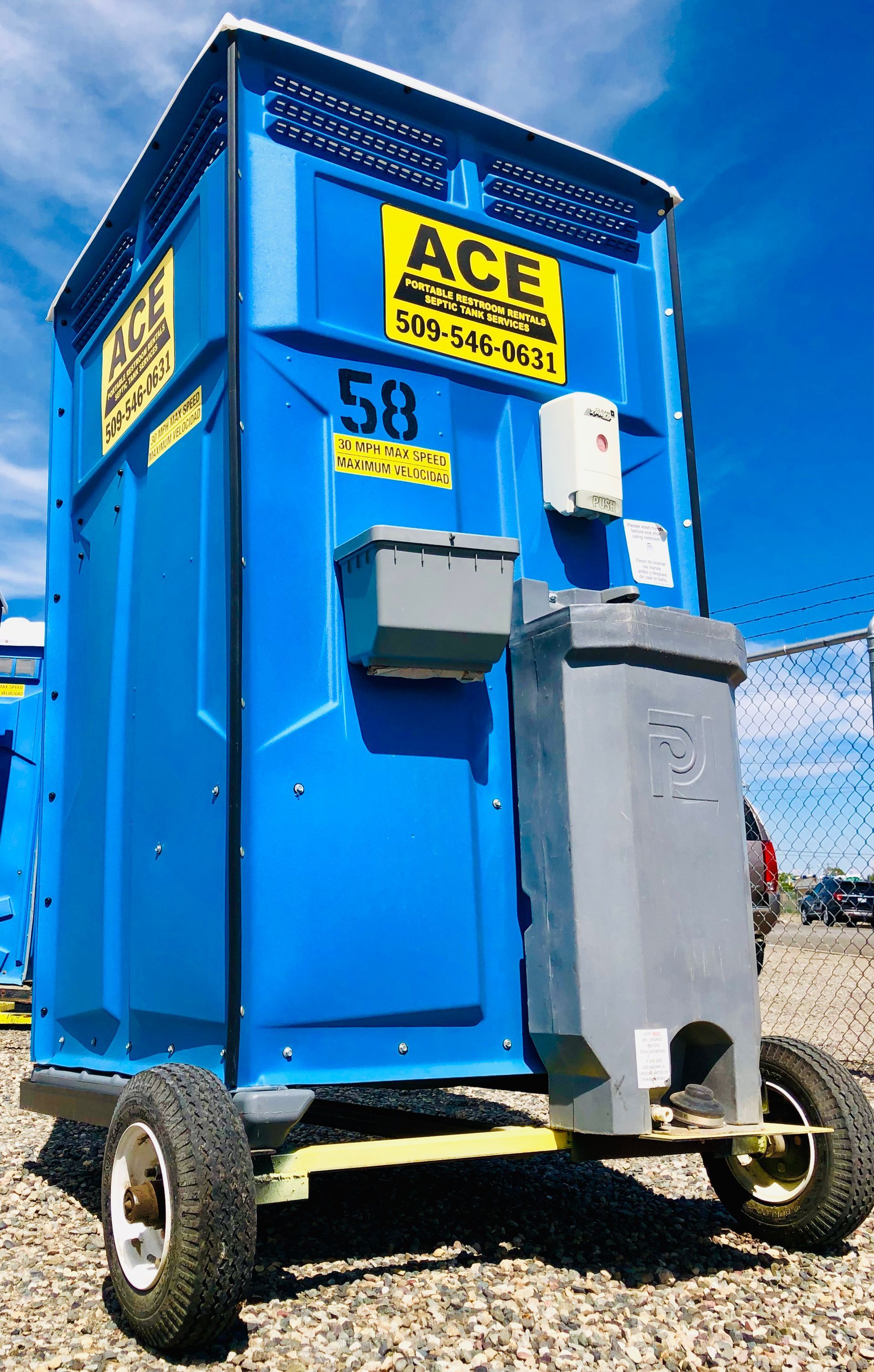 Blue portable toilet trailer with yellow ACE sign and gray utility boxes outdoors under a blue sky