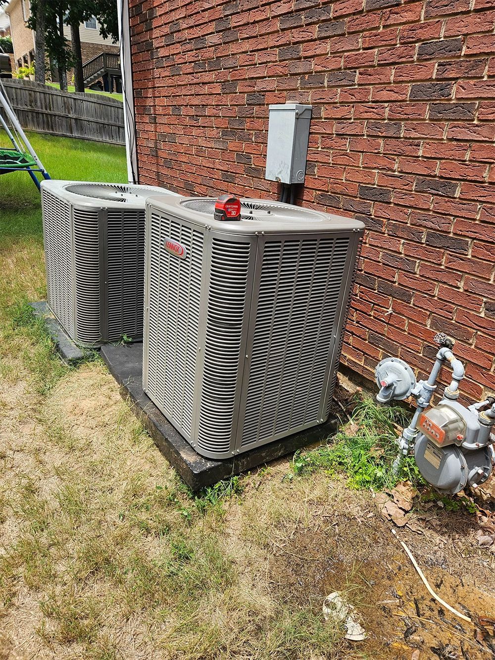 Two gray outdoor AC units beside a brick house wall on a grassy yard