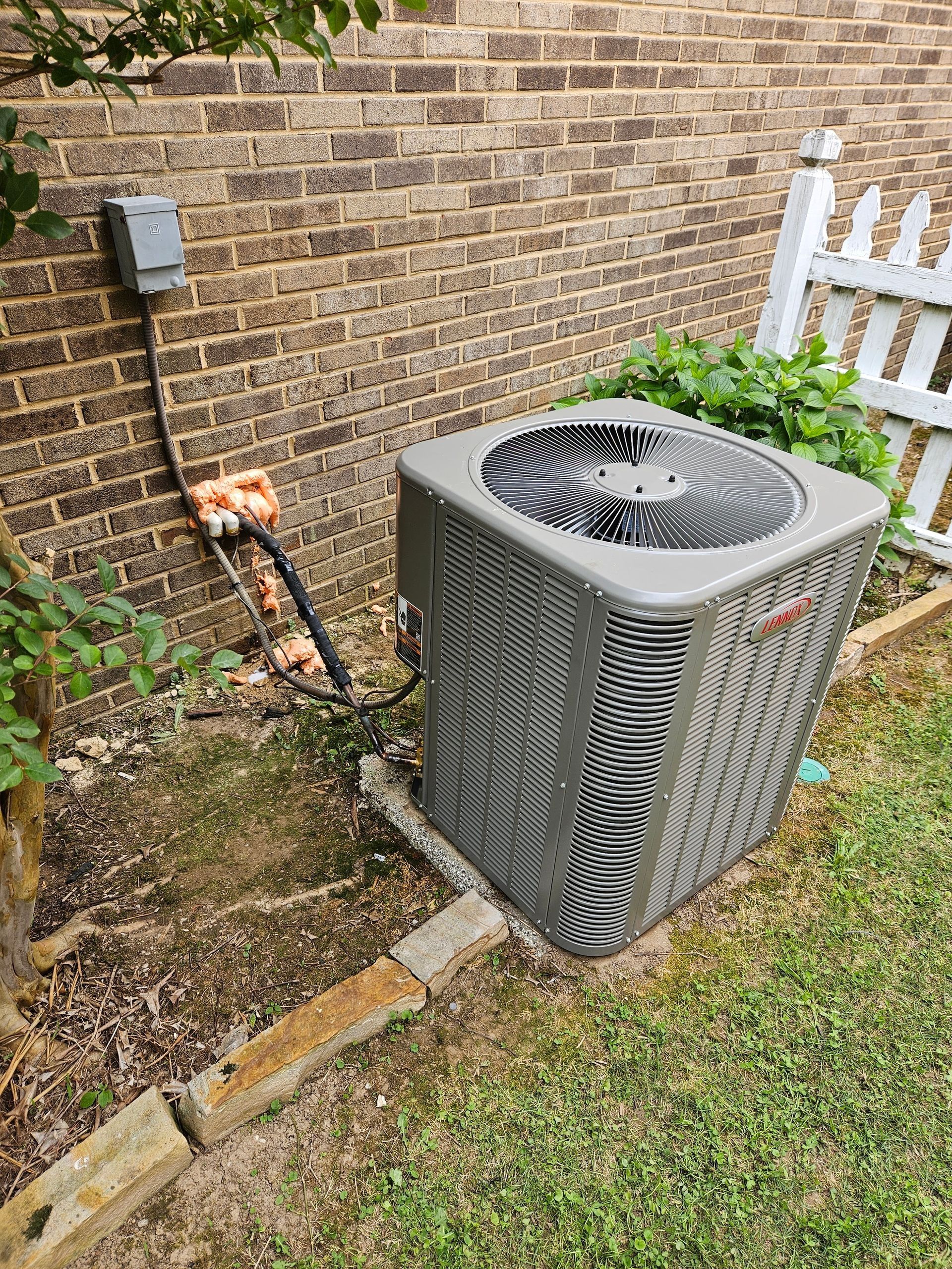 Outdoor air conditioner unit beside a brick wall with pipes, grass, and a white fence.