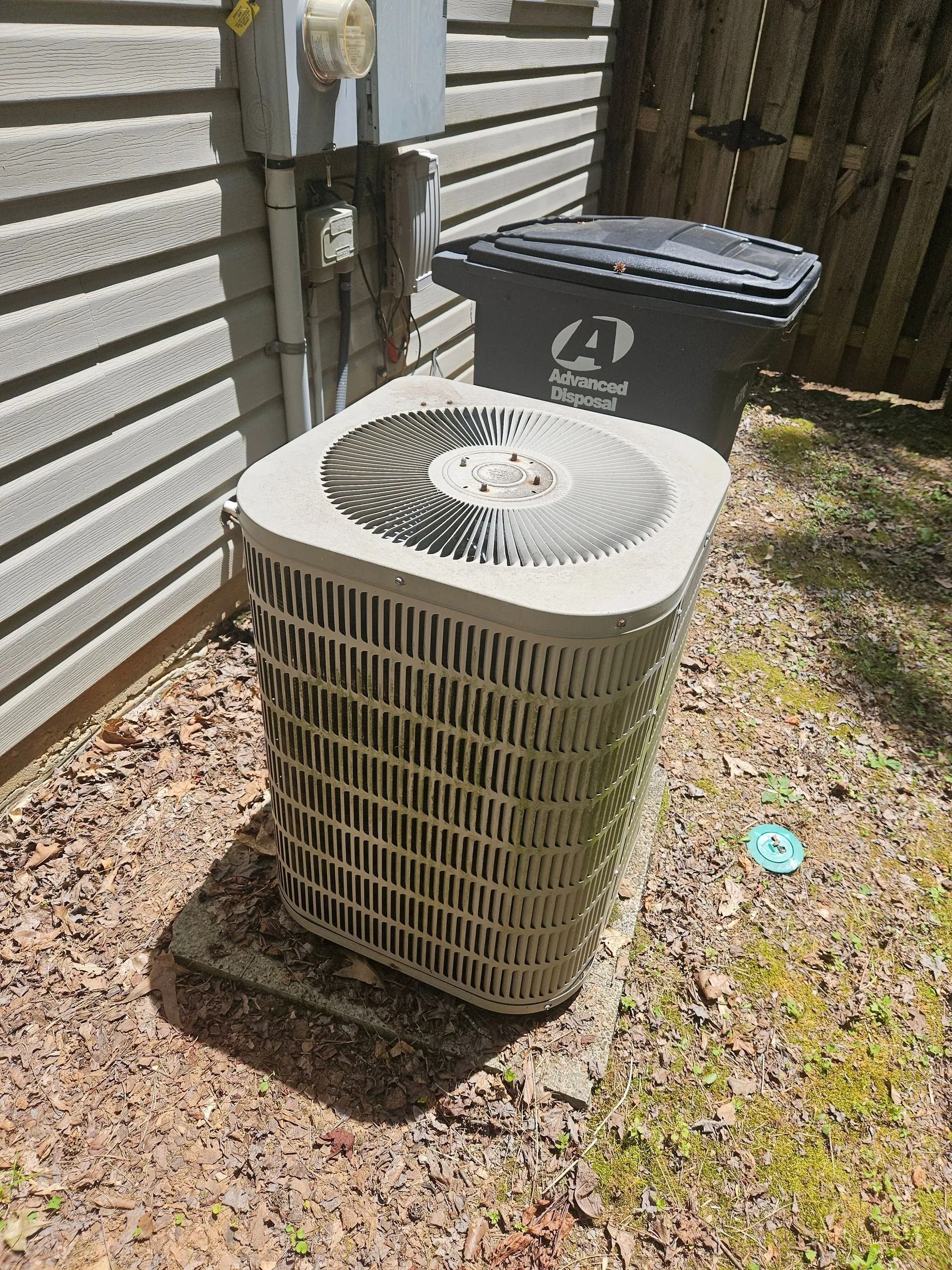 Outdoor air conditioner units beside a house wall and fence in sunlight.