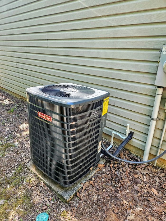 Outdoor air conditioner condenser beside house siding, with a hose and fallen leaves nearby