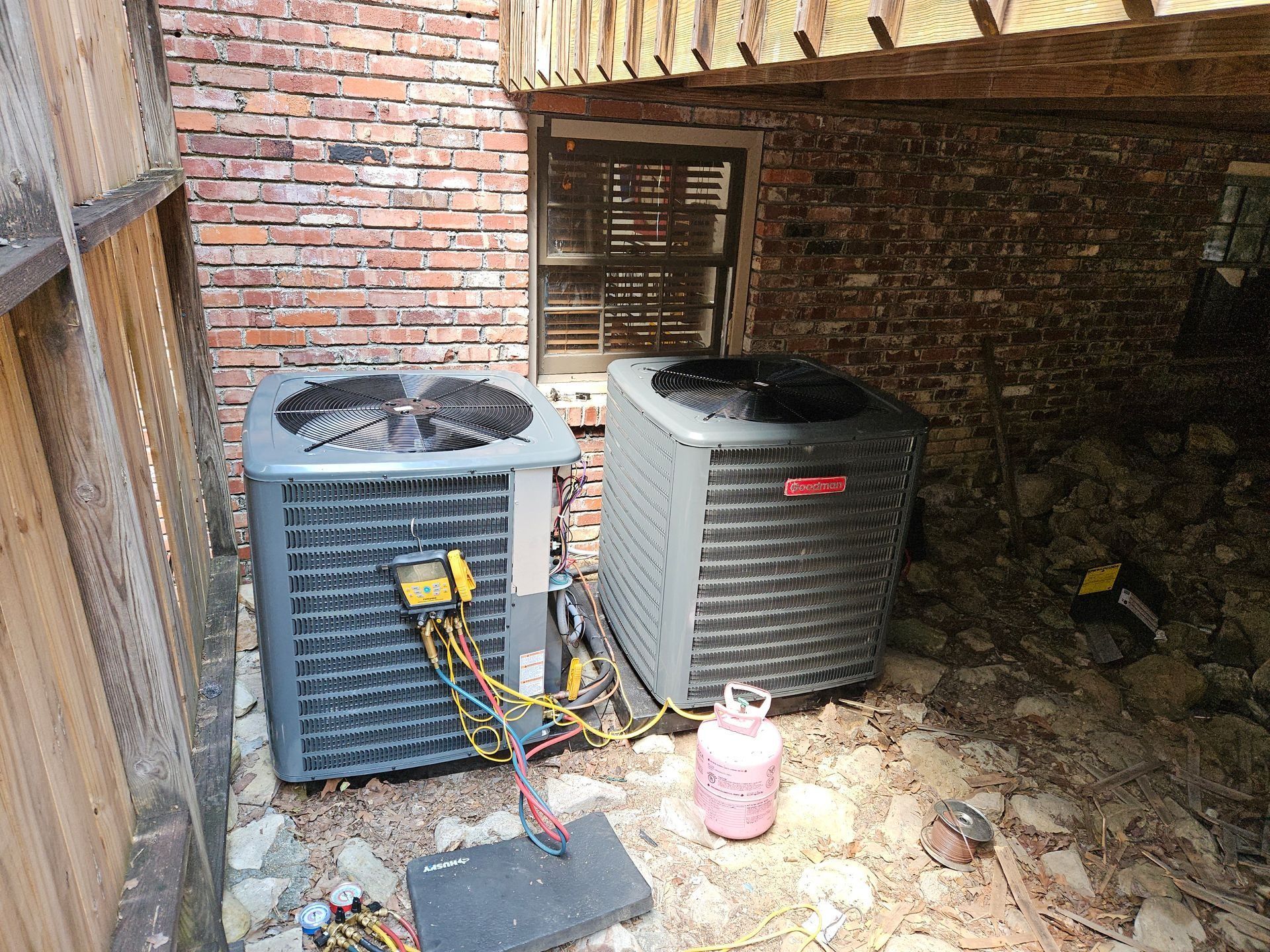 Two outdoor HVAC units beside a brick wall, with wiring and a propane tank on a cluttered ground.