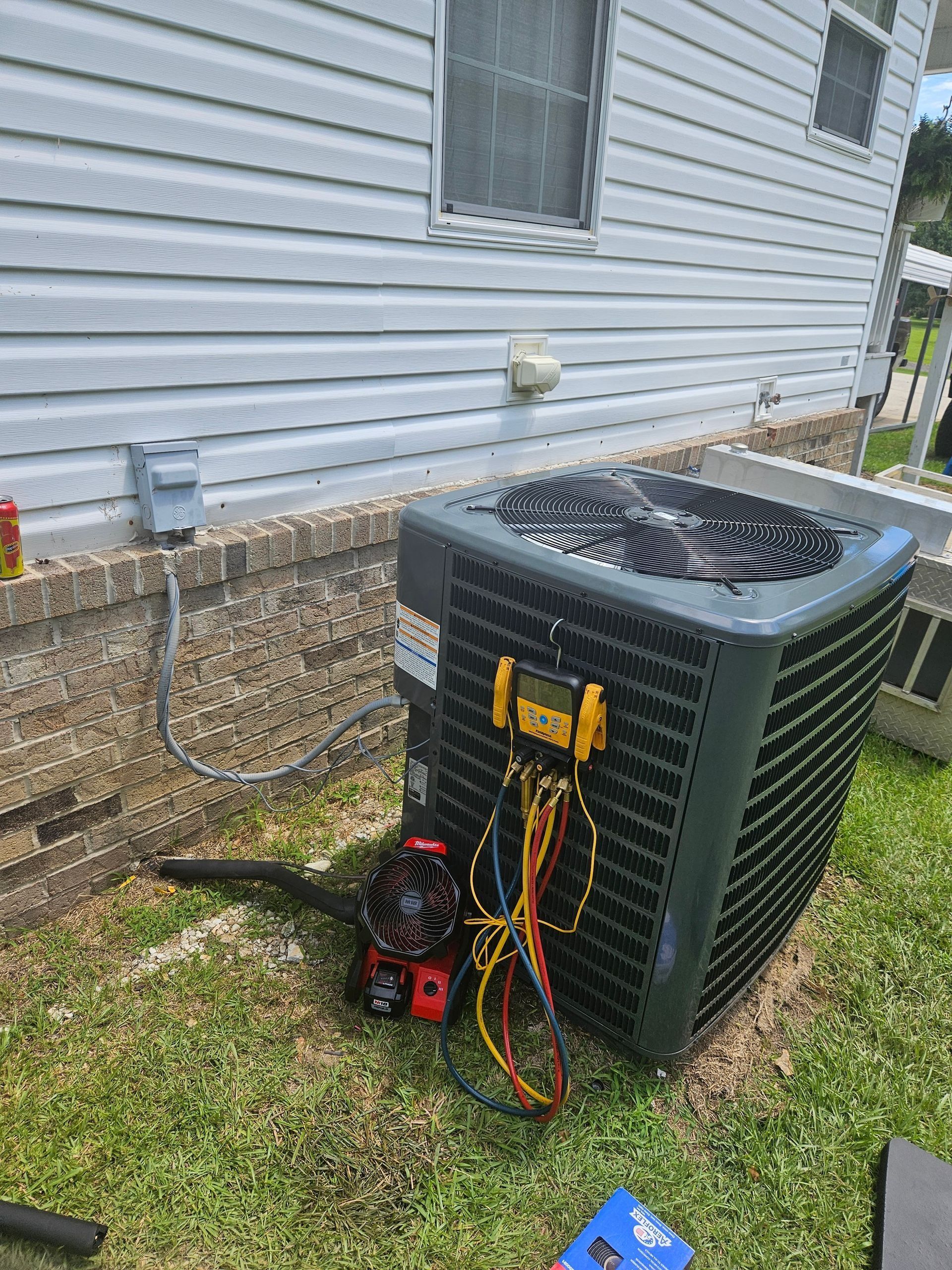 Outdoor air conditioner unit beside a house wall with tools and hoses attached, and a blue item on the grass.