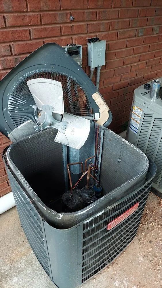 Damaged outdoor air conditioner with fan blades exposed against a brick wall