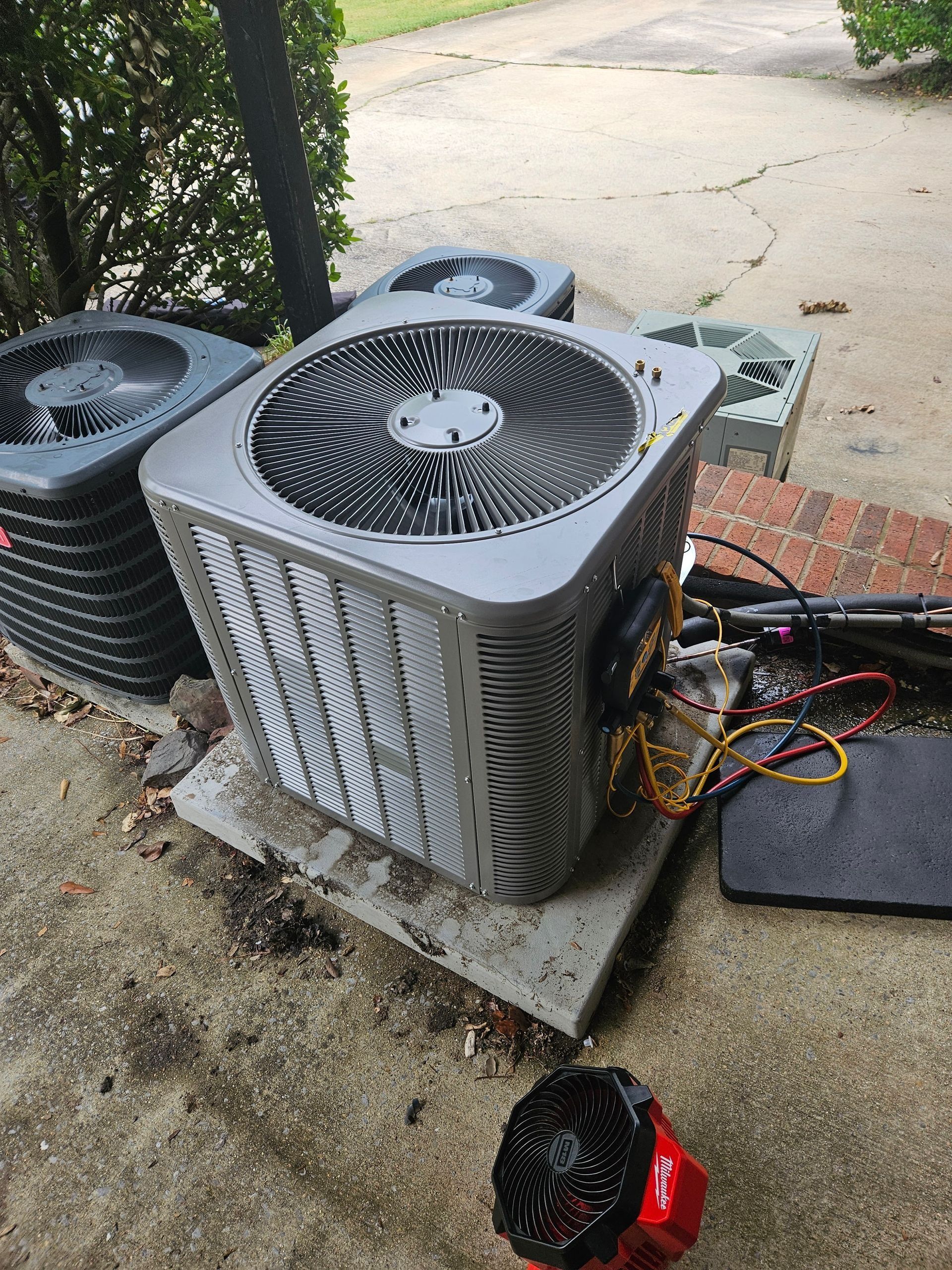Outdoor HVAC condenser units on a concrete pad with exposed electrical wiring and a work mat nearby.