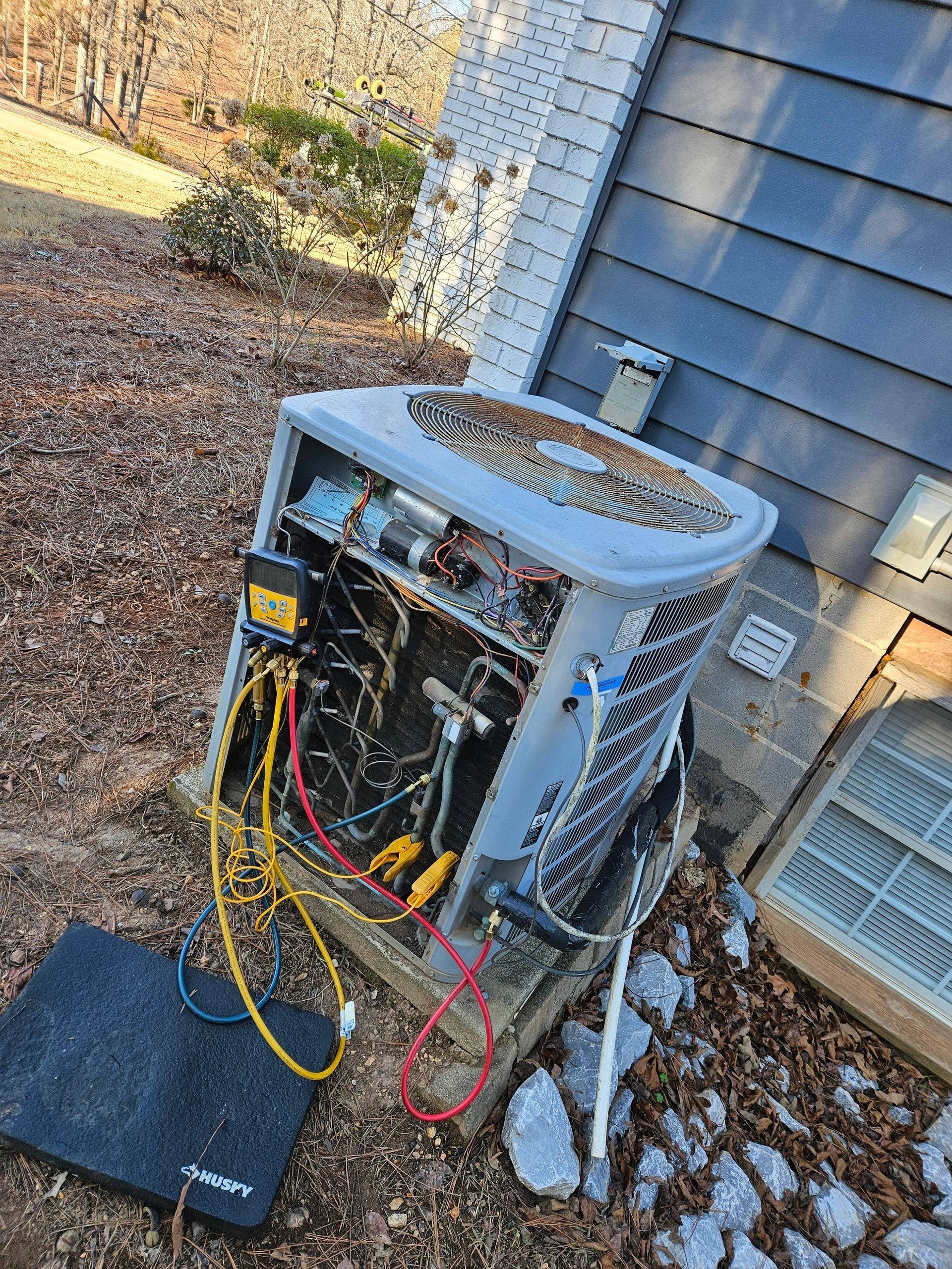Outdoor HVAC unit with its front panel open, showing exposed wiring beside a house wall.