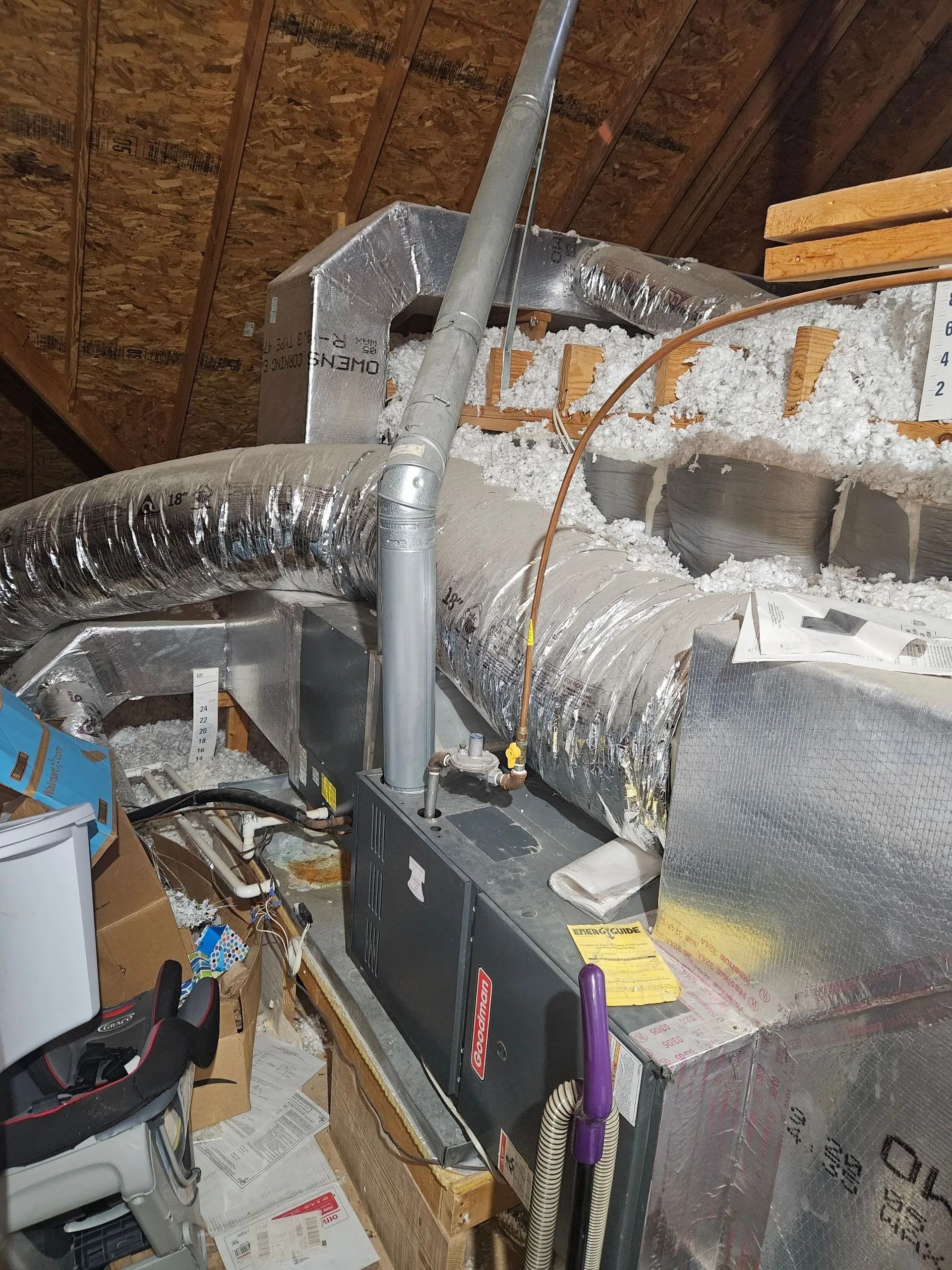Attic HVAC setup with silver ductwork, insulated pipes, and a furnace unit among storage items