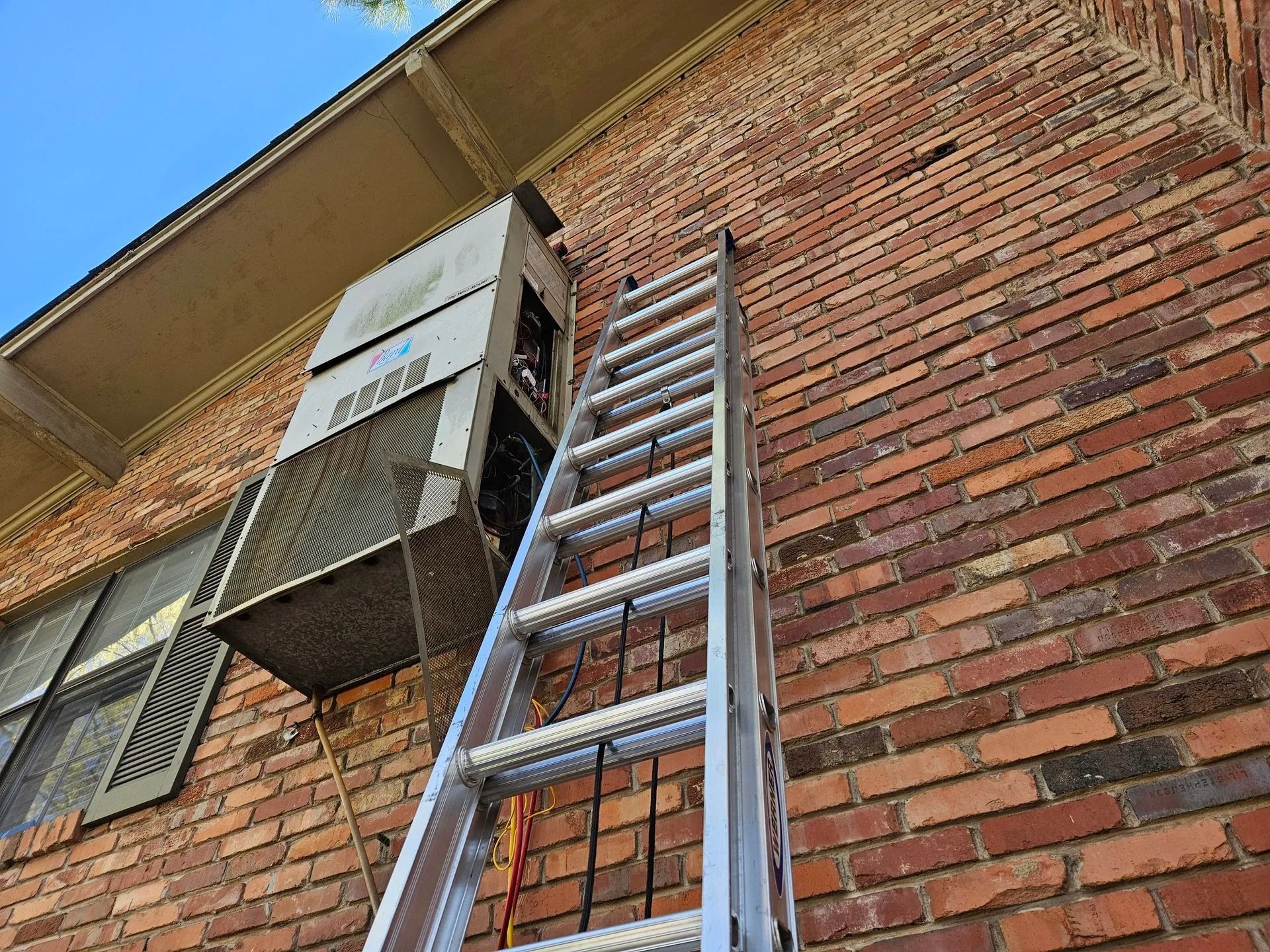 Aluminum ladder leaning against a brick building beneath a window air conditioner and awning