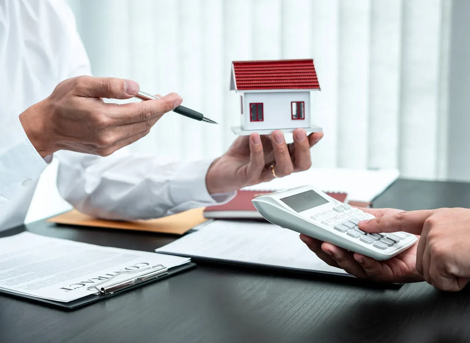 A man is holding a model house and a calculator while another man holds a pen.