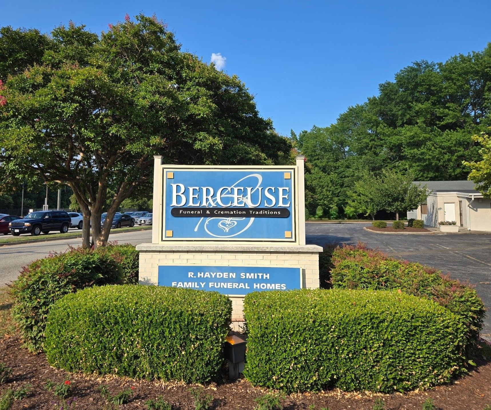 Sign for Berceuse Family Funeral Home; blue sign with white text, surrounded by green shrubs.