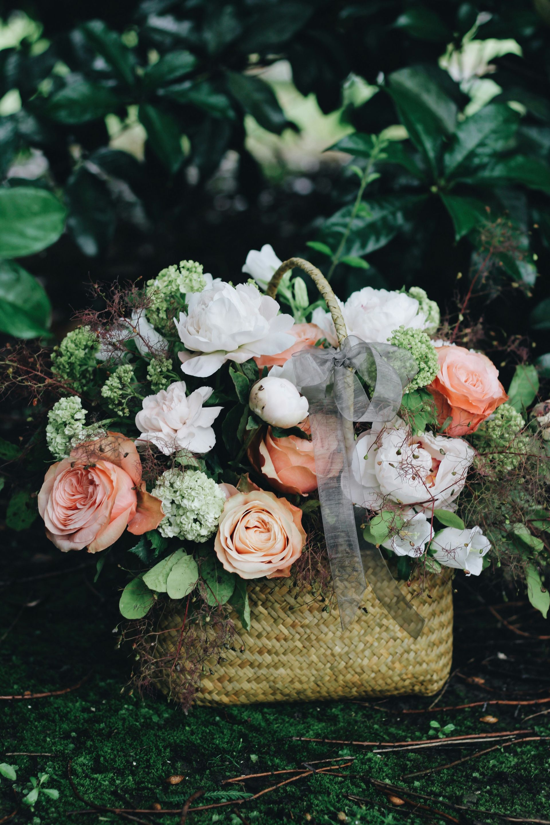Basket of pastel-colored flowers, accented with greenery and a gray ribbon, sits on moss.