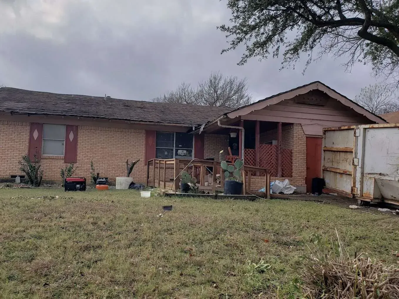 Single-story brick house with boarded windows, damaged front porch, and cloudy sky above.