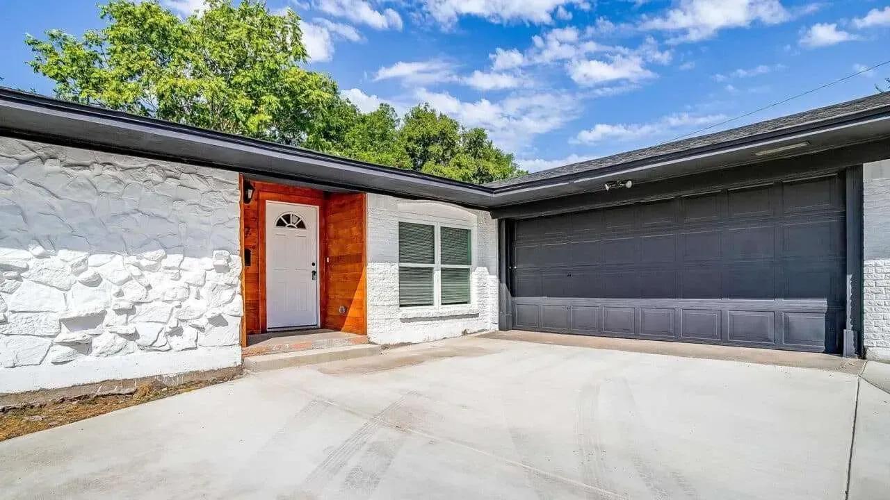Modern house exterior with white stone wall, orange door, and large gray garage door under a blue sky