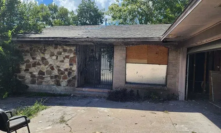 Abandoned house exterior with stone facade, boarded window, and broken driveway in daylight