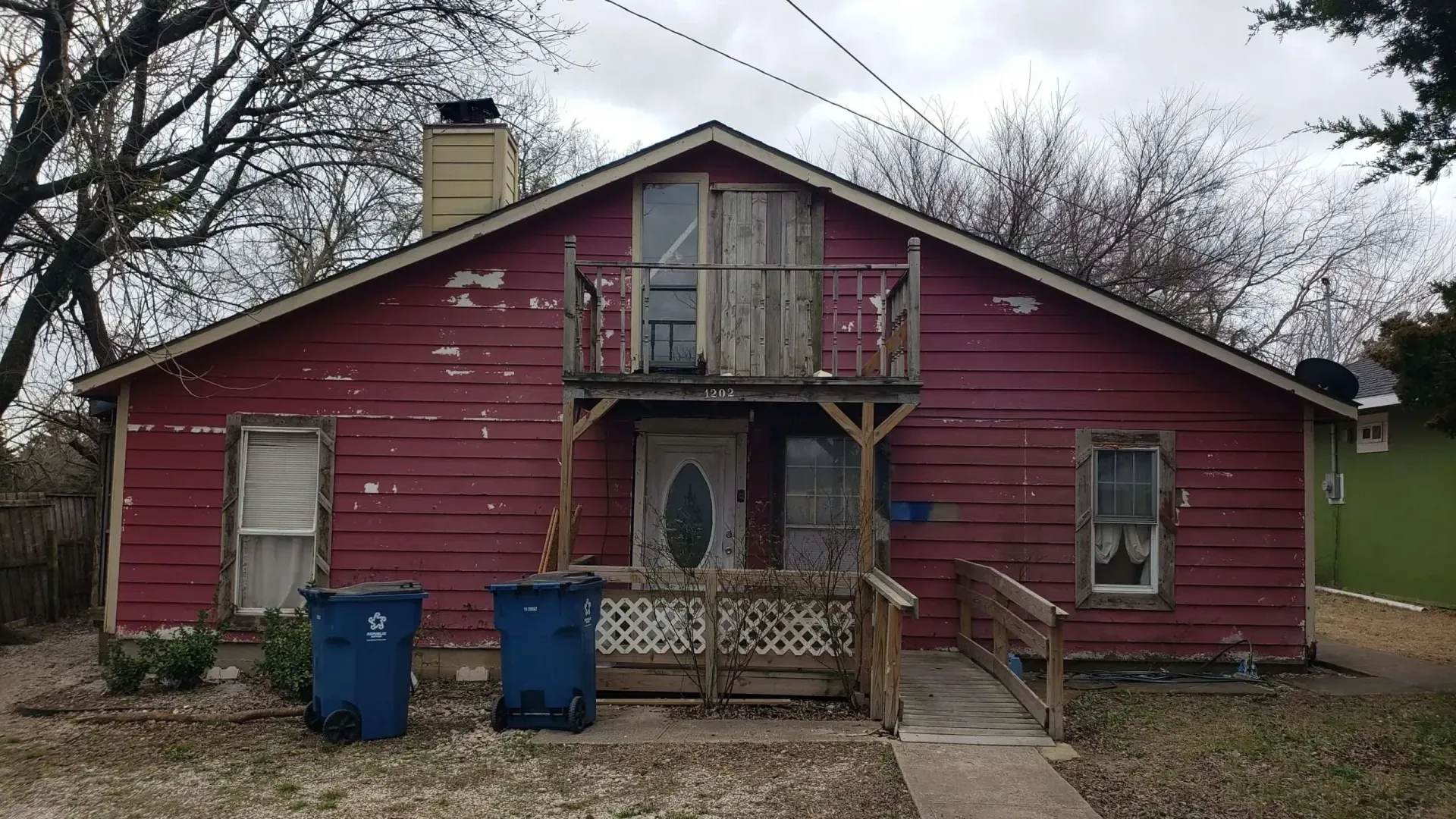 Weathered red house with a small front balcony and two blue bins in the yard