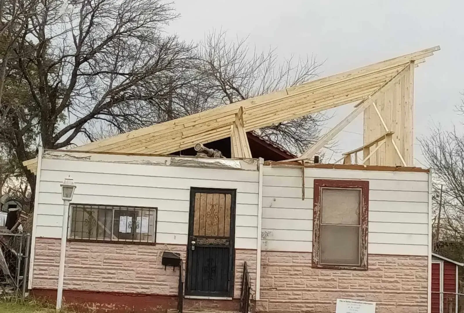 Damaged house with roof torn off, white siding, and bare trees in the background