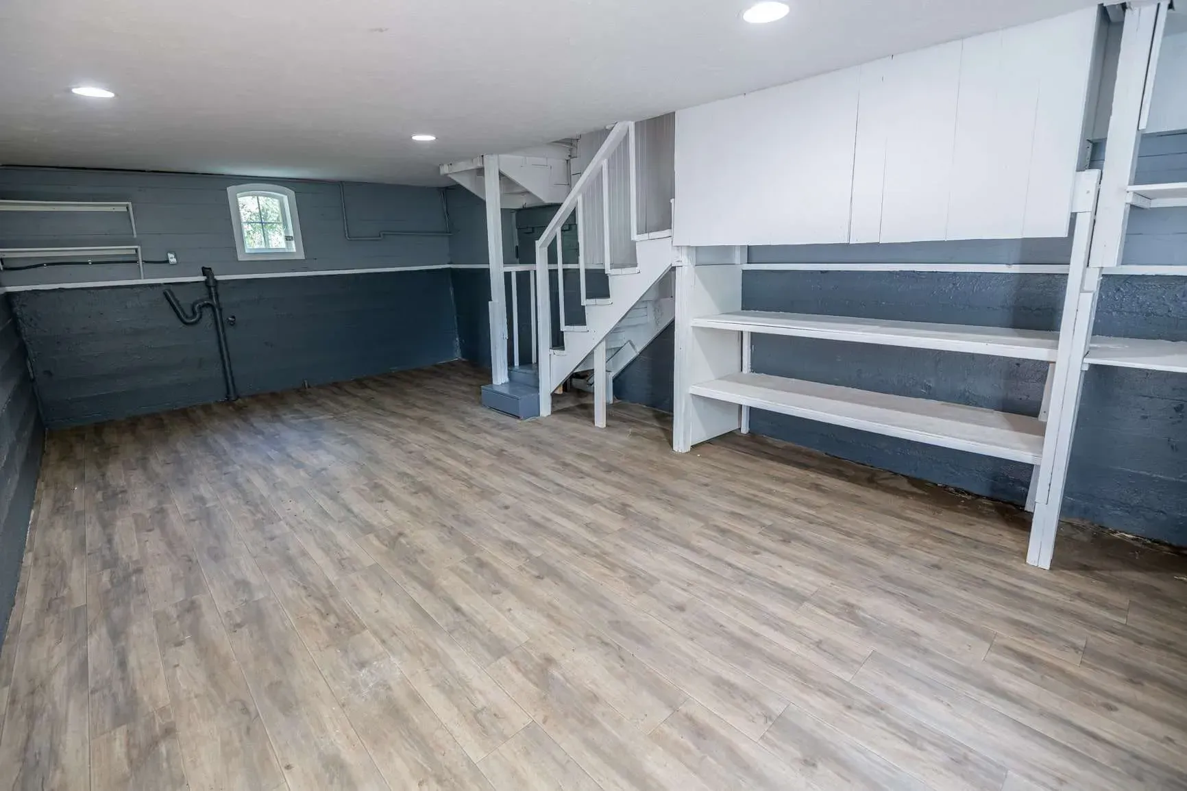 Empty finished basement with gray walls, wood-look floor, staircase, and built-in white shelving.