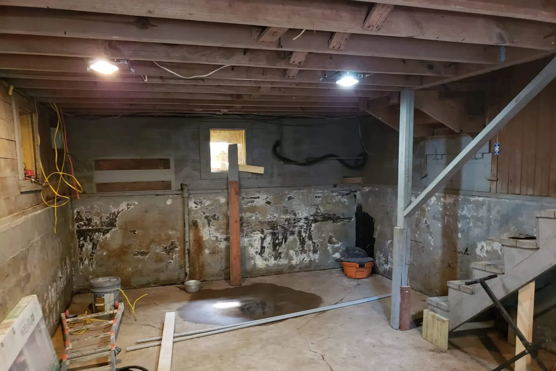 Unfinished basement with exposed beams, concrete walls, and scattered tools and debris