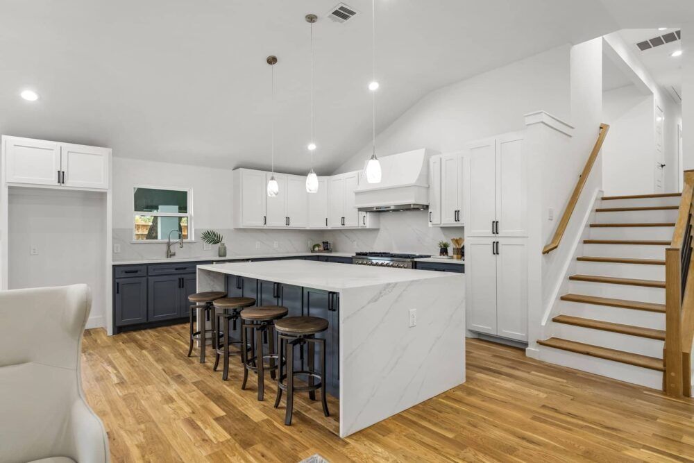 Bright modern kitchen with white island, dark stools, wood floors, and staircase on the right