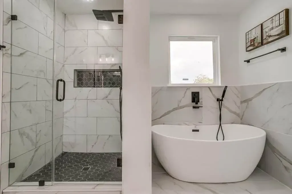 Modern bathroom with glass shower, freestanding tub, marble tile, and small window.