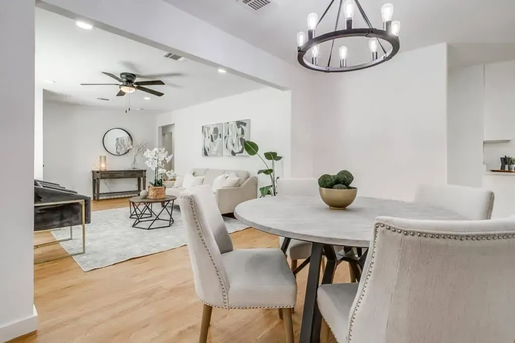 Bright open dining room with round table, upholstered chairs, and a glimpse of a living area beyond.
