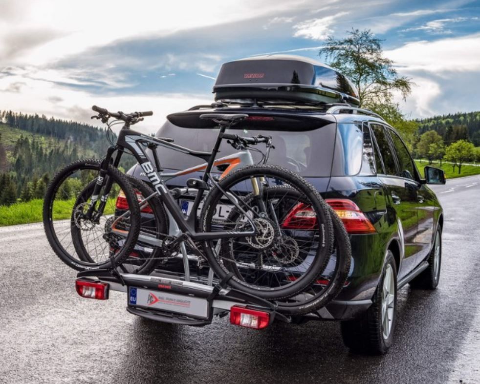 SUV nero con box portatutto sul tetto e due biciclette sul portapacchi posteriore parcheggiato su una strada di montagna bagnata.