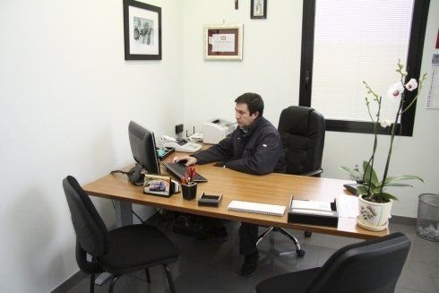 Persona seduta a una scrivania in un piccolo ufficio, al lavoro al computer, con documenti e una pianta in vaso nelle vicinanze.