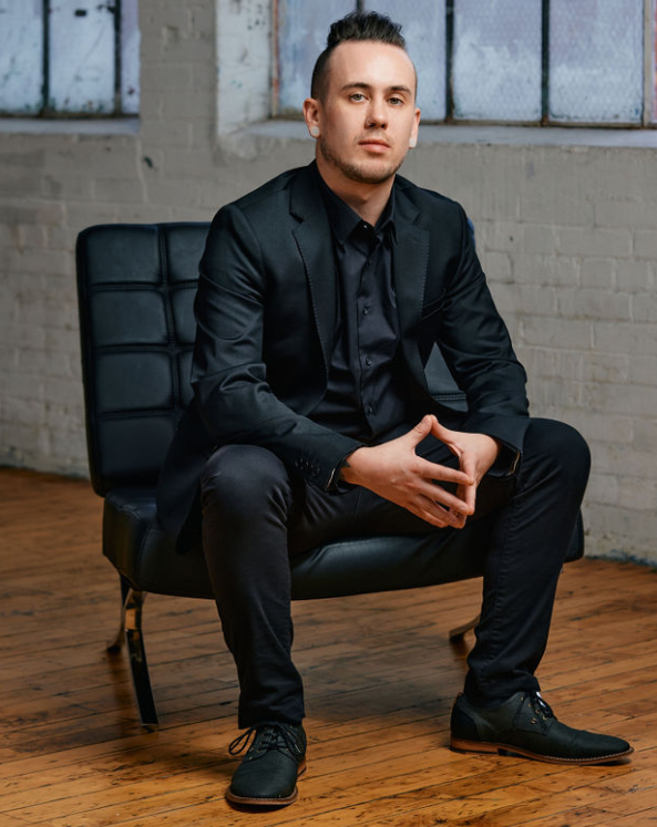 Man in black suit sitting on a modern chair in a loft-style room with brick walls and wooden floor